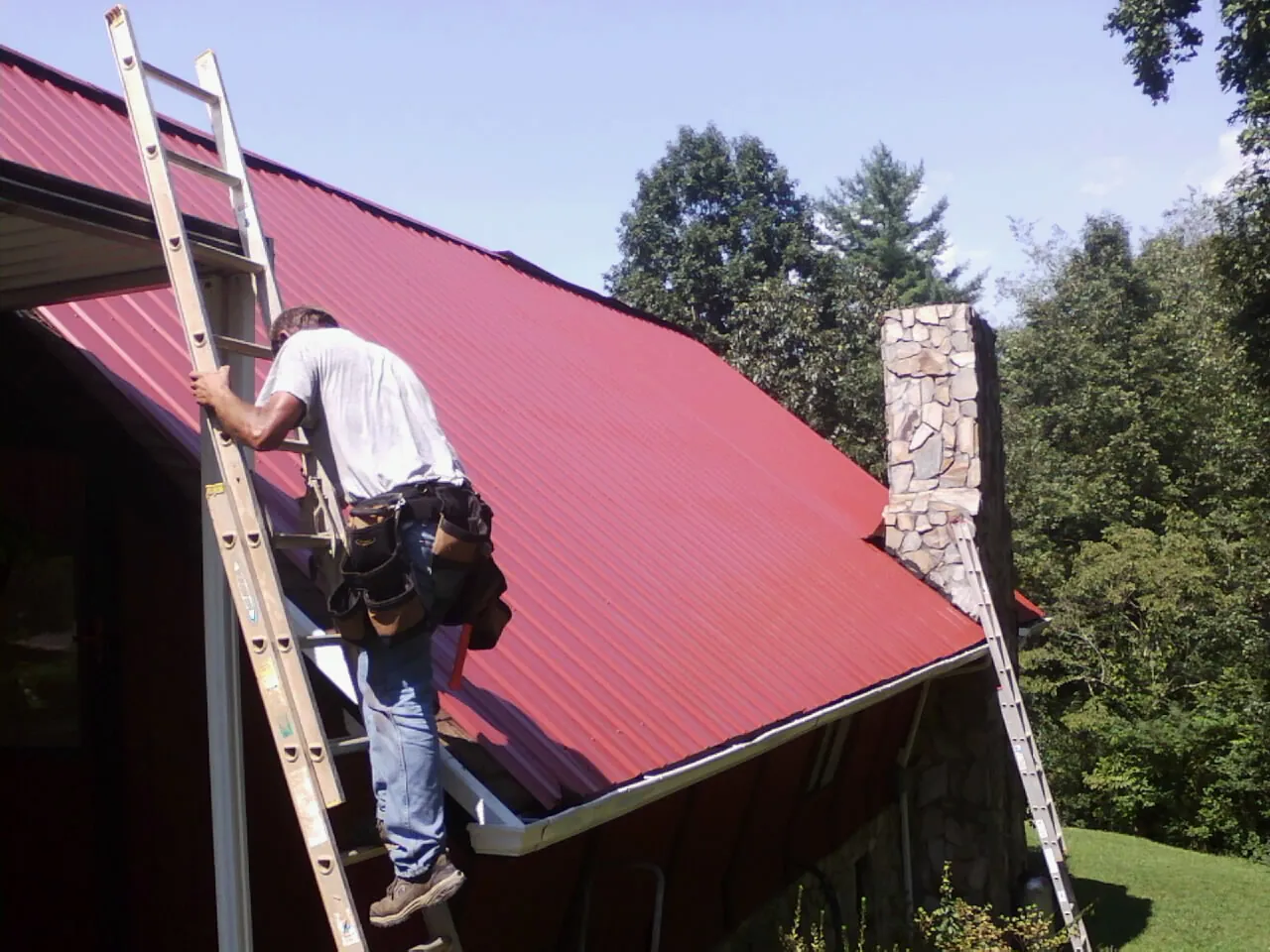 Roofer on a ladder beside a red metal roof and stone chimney — Soffit & Fascia Repair by Roofing Brothers in Stony Creek